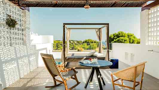 En terrasse med to stoler og et lite bord samt en dagseng med utsikt over vegetasjon og hav. (AI-generert tekst)