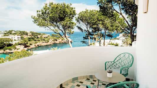 En balkong med to stoler og et bord, utsikt over havet og landskap med furutrær. (AI-generert tekst)