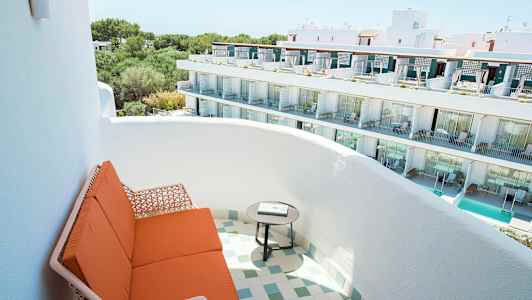 Bildet viser en balkong med oransje sittemøbler og et rundt bord, med utsikt over en hvit bygning med flere etasjer og balkonger. (AI-generert tekst)