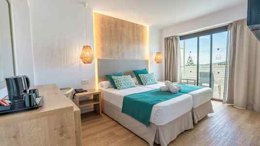 Et hotellrom med to enkeltsenger, en trevegg bak sengen, turkise laken og puter, og en balkong med utsikt. (AI-generert tekst)