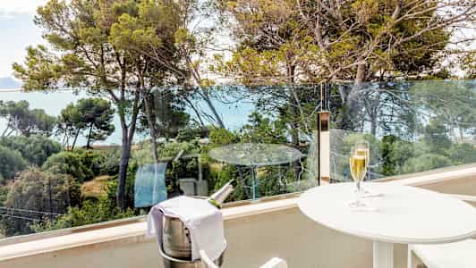 En balkong med utsikt over trær og havet. På balkongen står et hvitt bord med et glass champagne og en bøtteholder med en champagneflaske. (AI-generert tekst)