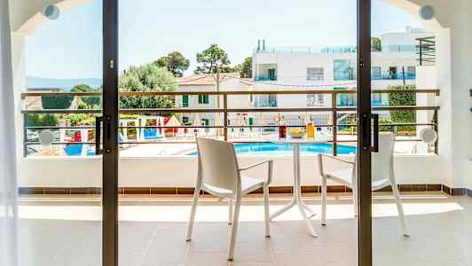 En balkong med to stoler og et bord med drikke og snacks. Utsikt over et bassengområde og omkringliggende bygninger. (AI-generert tekst)