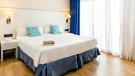 Et hotellrom med en bred seng, hvitt sengegavl, blå puter og gardiner samt et lyst treparkettgulv. (AI-generert tekst)