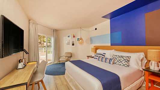 Bilde av et hotellrom med moderne interiør, en seng med blått sengeteppe, skrivebord og stol samt en balkongdør. (AI-generert tekst)