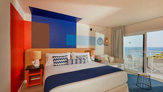 Et rom med en dobbeltseng, dekorative malte vegger, sittemøbler og utsikt mot havet gjennom store vinduer. (AI-generert tekst)