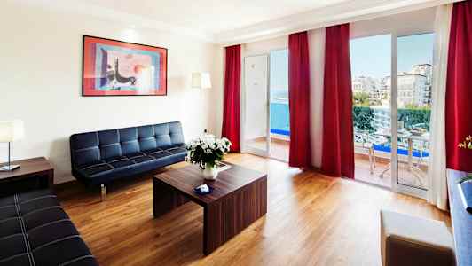 En moderne stue med utsikt mot havet gjennom store vinduer og balkong, møblert med svarte sofaer og et trebord. (AI-generert tekst)