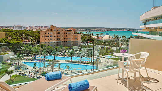 Utsikt fra en balkong over et basseng og hage med palmer, hotellbygninger og hav i bakgrunnen. (AI-generert tekst)