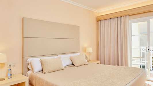 Rom med en dobbeltseng, beige sengetøy og lampe på nattbordet ved siden av vindu med gardiner. (AI-generert tekst)