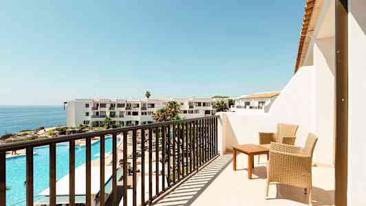 En balkong med to flettede stoler og et bord, utsikt over et hotellområde med utendørsbasseng og havet i bakgrunnen. (AI-generert tekst)