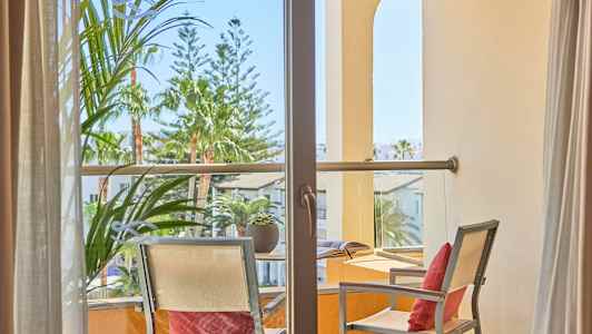 Utsikt gjennom en balkong med to stoler og et bord, omgitt av planter og bygninger. (AI-generert tekst)