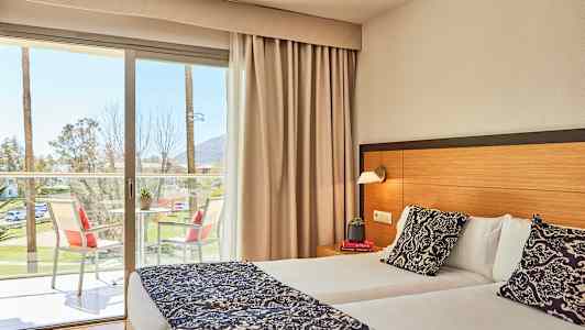 Bilde av et hotellrom med en dobbeltseng, et bord med stoler på balkongen og utsikt mot palmer utenfor. (AI-generert tekst)