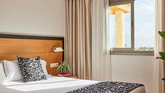 Hotellrom med seng, nattbord og et vindu som slipper inn naturlig lys. (AI-generert tekst)