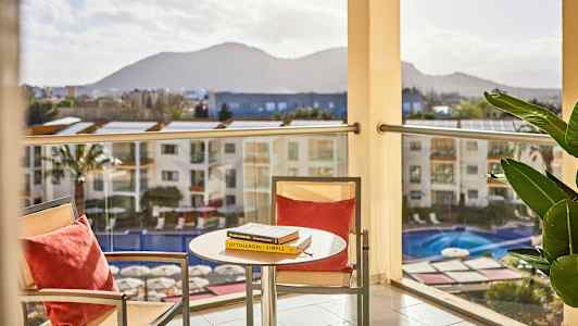 En utsikt fra en balkong med et bord og to stoler i forgrunnen samt et basseng nedenfor. Fjell kan sees i bakgrunnen. (AI-generert tekst)