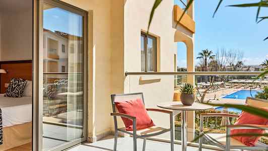 En balkong med to stoler og et bord med en plante, utsikt over et utendørsbasseng og omkringliggende hage. Et rom med seng er synlig i bakgrunnen. (AI-generert tekst)