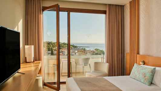 Et hotellrom med en seng, skrivebord, TV og en åpen balkongdør som leder til en balkong med utsikt over havet og trær. (AI-generert tekst)
