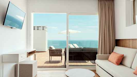 Et lyst rom med en sofa og bord, som har en utendørs balkong med sittemøbler og utsikt over havet. (AI-generert tekst)