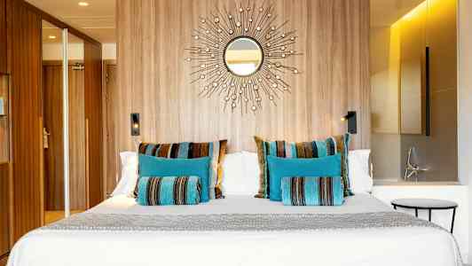 Et hotellrom med en stor seng dekorert med fargerike puter i blått og stripete mønster, en trevegg med et speilmotiv og moderne belysning. (AI-generert tekst)