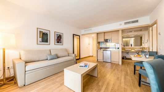 Et moderne hotellrom med en beige sofa og et bord på tregulv, kjøkkenkrok og bad i bakgrunnen. (AI-generert tekst)