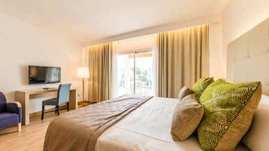 Et hotellrom med en stor seng, grønne puter, et bord, en stol og en TV. Vinduer med gardiner slipper inn naturlig lys. (AI-generert tekst)