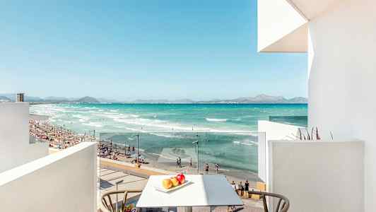 Utsikt fra en balkong mot en strand med turkist vann, sand og mennesker som bader. (AI-generert tekst)