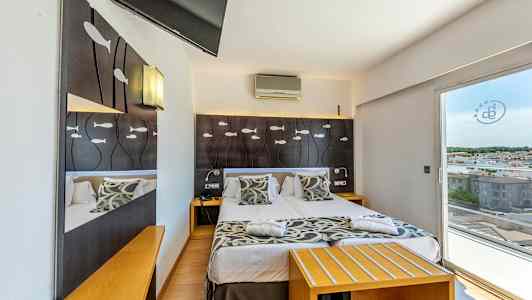 Hotellrom med dobbeltseng, dekorert med fiskemotiv på sengegavlen og utsikt mot byen gjennom glassdører. (AI-generert tekst)