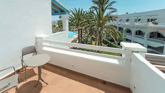 En balkong med bord og stoler, med utsikt over palmer og et basseng. (AI-generert tekst)