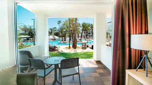 Utsikt fra et rom med balkong mot et solfylt bassengområde. (AI-generert tekst)