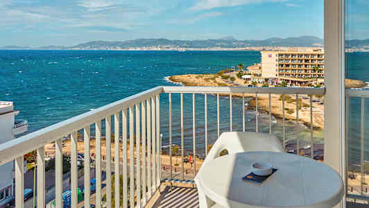 Utsikt fra en balkong mot en kyst med hav og bygninger i bakgrunnen. (AI-generert tekst)