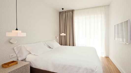 Rom med en seng, hvite laken og gardiner, samt veggmontert TV. (AI-generert tekst)