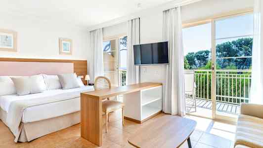 Et hotellrom med dobbeltseng, skrivebord og TV. Store glassdører fører ut til en balkong. (AI-generert tekst)