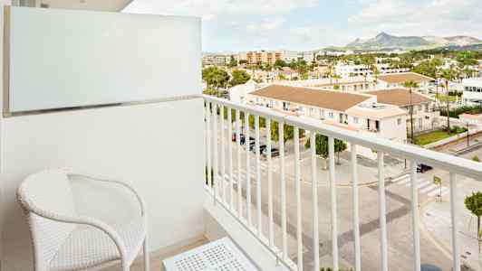 En balkong med to hvite stoler og et bord, med utsikt over en gate og bygninger. (AI-generert tekst)