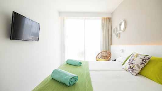 Et lyst hotellrom med en dobbeltseng, fargerike puter, sammenrullede håndklær og en flatskjerm-TV på veggen. (AI-generert tekst)