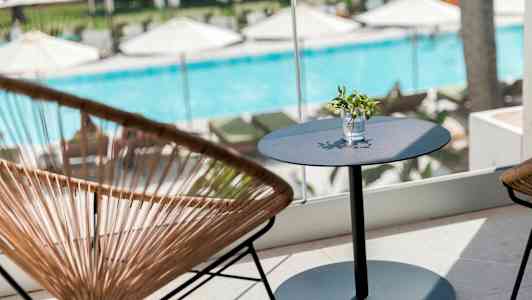 En sittegruppe med et bord og stoler foran et vindu med utsikt over et basseng og solsenger. (AI-generert tekst)