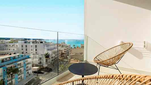 En balkong med to stoler i rotting og et rundt bord, med utsikt mot havet og byens bygninger. (AI-generert tekst)