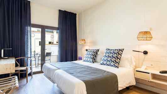 Et hotellrom med en dobbeltseng, blåe pynteputer og nattlamper. Et skrivebord med stol står ved siden av, samt en balkong med bord og stoler. (AI-generert tekst)