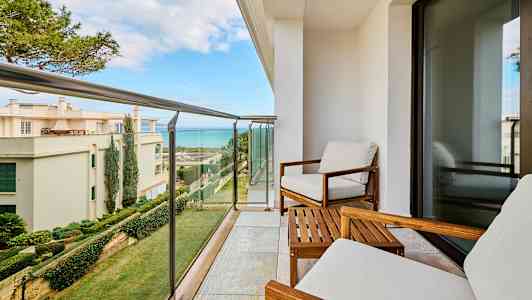 Balkong med to stoler, et bord og utsikt over boliger og hav. (AI-generert tekst)