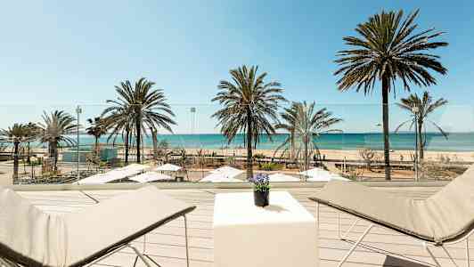 Utsikt fra en terrasse med solsenger og bord over en strand med palmer og hav. (AI-generert tekst)