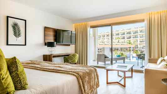 Et hotellrom med en seng, sofagruppe og balkong med utsikt over bassengområdet og hotellet. (AI-generert tekst)