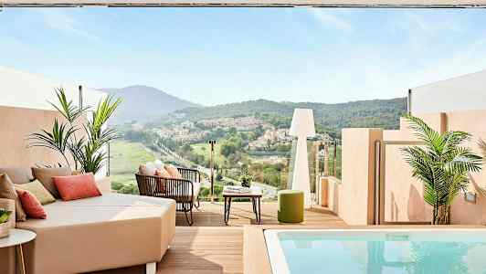 En terrasse med utsikt over en grønn dal og hus i det fjerne, med sittemøbler og et lite basseng. (AI-generert tekst)