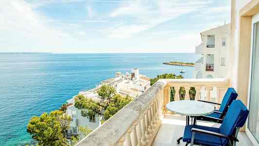 Utsikt fra en balkong med bord og stoler over havet og omkringliggende bygninger. (AI-generert tekst)