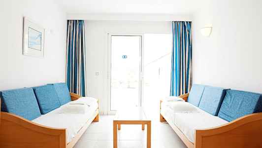 To sofaer med blå puter står overfor hverandre, med et trebord imellom og blåhvite gardiner ved vinduet i bakgrunnen. (AI-generert tekst)