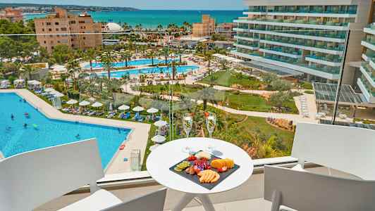 Utsikt fra en balkong mot et hotellbasseng og havet, med en fruktfat og to glass på et bord. (AI-generert tekst)