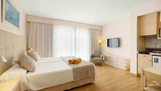 Et hotellrom med dobbeltseng, to håndklær på sengen, en lenestol, en veggmontert TV og et lite kjøkkenhjørne. (AI-generert tekst)