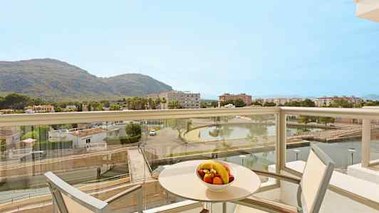 En balkong med utsikt over et landskap som inkluderer fjell, bygninger og en dam. På bordet er en bolle med frukt. (AI-generert tekst)