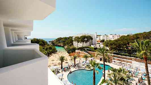 Utsikt fra en balkong mot et hotellområde med basseng, strand og trær. (AI-generert tekst)