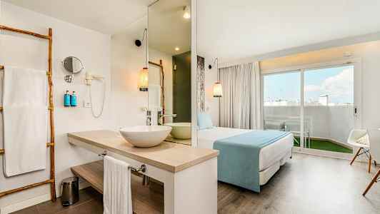 Hotellrom med bad, sovesone og sittegruppe. Stor vindusdør mot en utendørs terrasse. (AI-generert tekst)