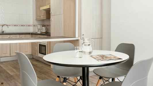 En spisestue med et rundt bord, grå stoler og et tilstøtende kjøkkenområde med tremøbler og hvitvarer. (AI-generert tekst)