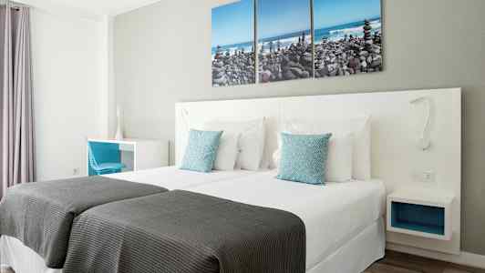 Et hotellrom med to enkeltsenger, hvitt sengetøy og blå pynteputer. På veggen henger et tredelt maleri som viser en steinstrand. (AI-generert tekst)