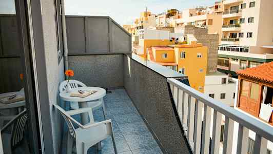 En balkong med utsikt over omkringliggende bygninger. Det er et bord med to stoler, en bok og en blomst på bordet. (AI-generert tekst)