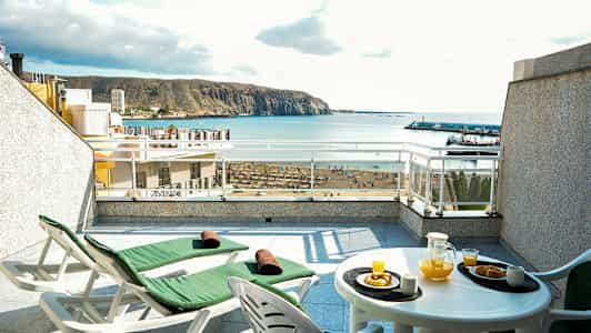 Terrasse med to solsenger og et bord med appelsinjuice og bakverk, havutsikt i bakgrunnen. (AI-generert tekst)